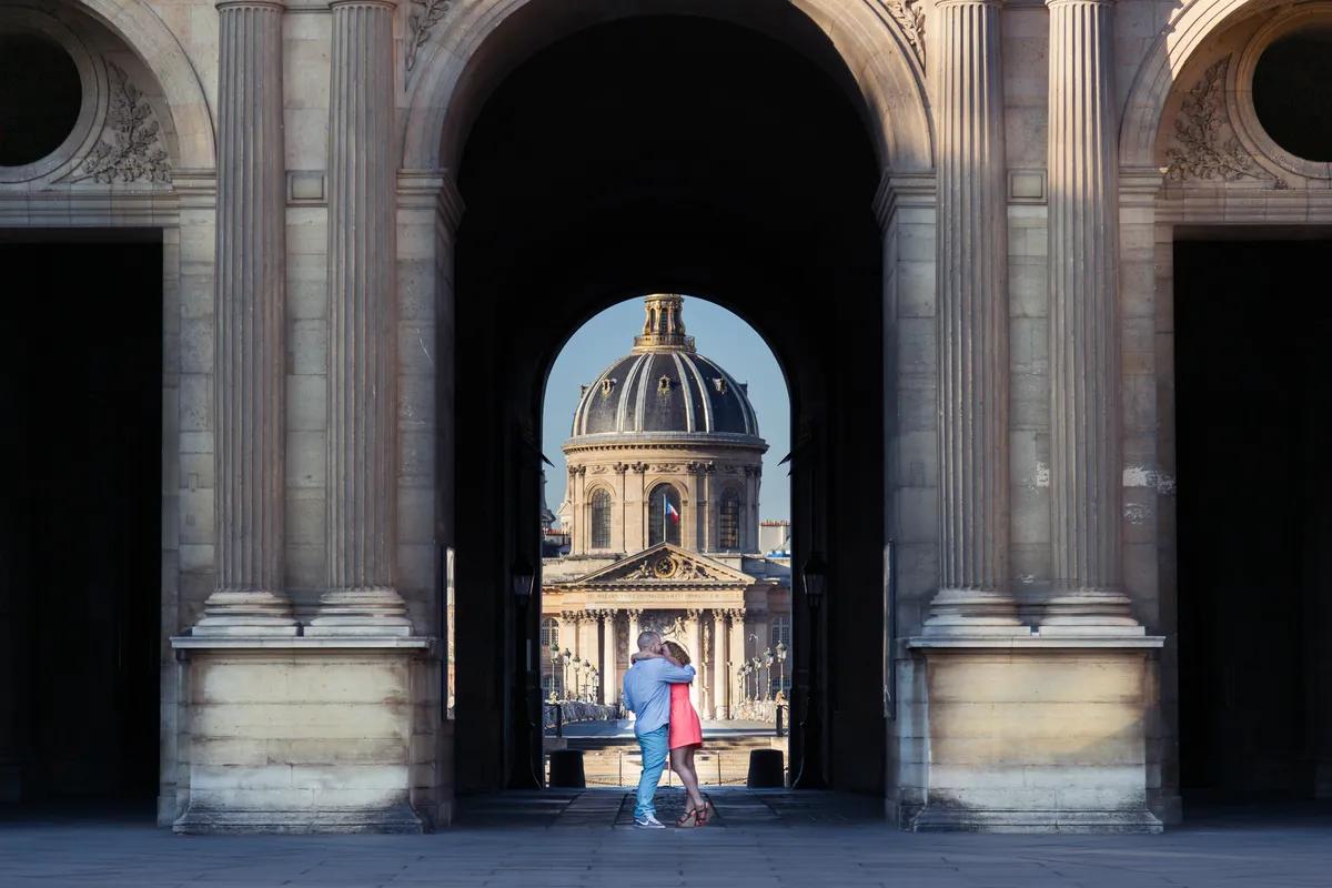 Couple marchant sous une arche avec vue sur l’Institut de France lors d’une séance photo à Paris.
