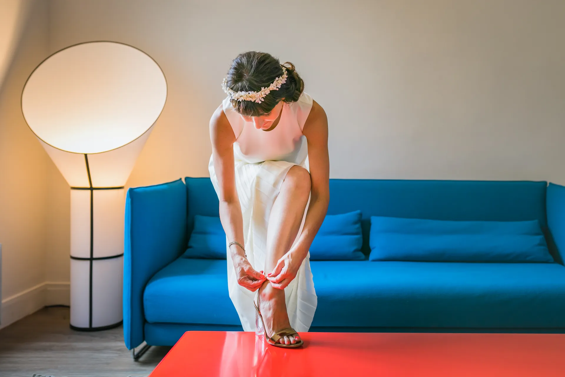 La mariée, une couronne de fleurs dans les cheveux, attache sa chaussure devant un canapé bleu pendant les préparatifs à Paris.