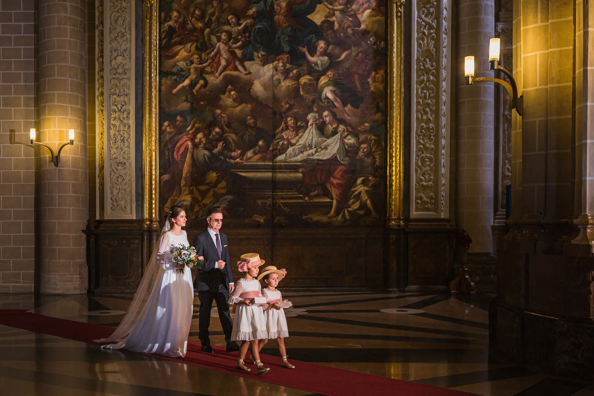 La mariée avance dans l'église avec son père et deux petites filles en robe blanche, sur un tapis rouge.