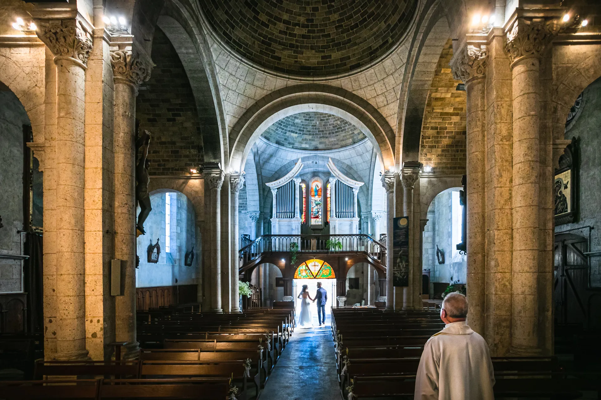 Mariés main dans la main sortant d'une église de Bordeaux, silhouette rétroéclairée sous les voûtes de pierre