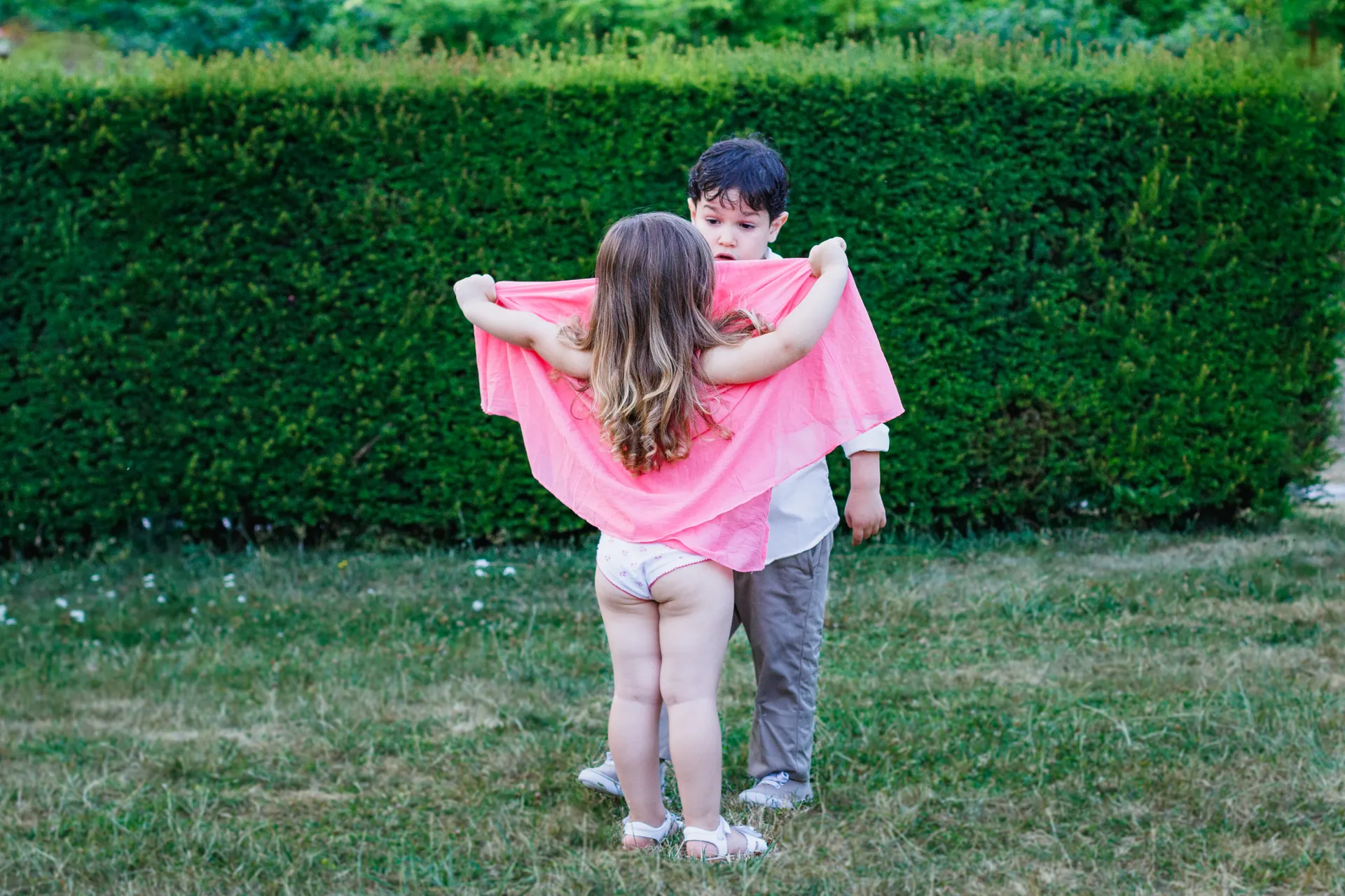 Deux enfants jouant avec un tissu rose comme une cape, de dos devant une haie verdoyante au Château de Vallery