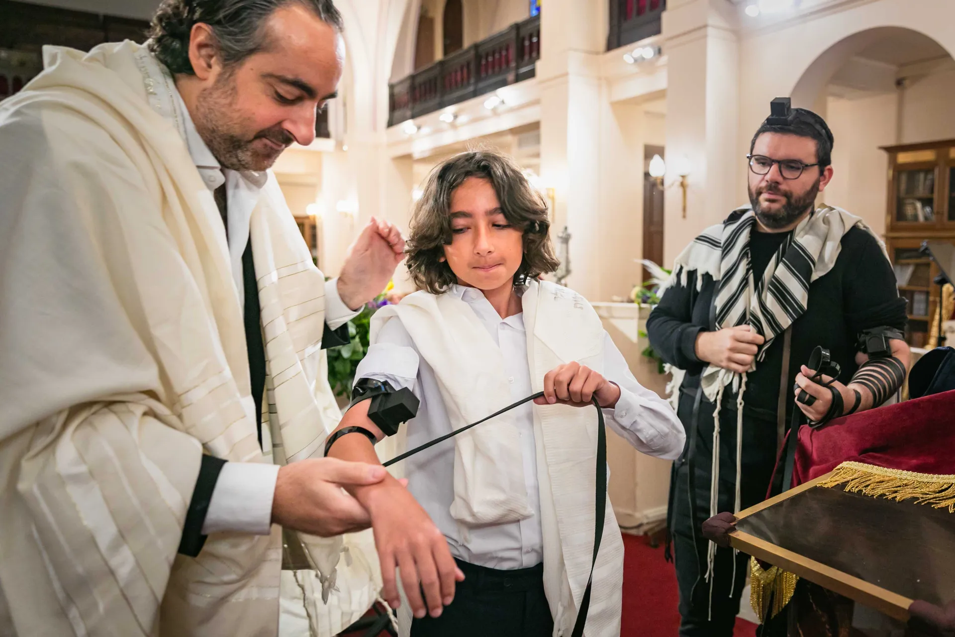 Un homme aidant le jeune garçon à poser les tefillin, maintenus par les lanières de cuir (retzuot), lors de sa Bar-Mitzvah dans une synagogue à Paris